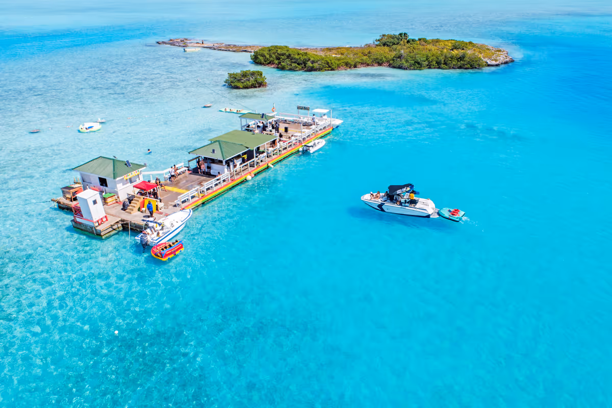 Aerial view of a floating bar with green roofs moored on turquoise water, boats alongside and a small green cay behind
