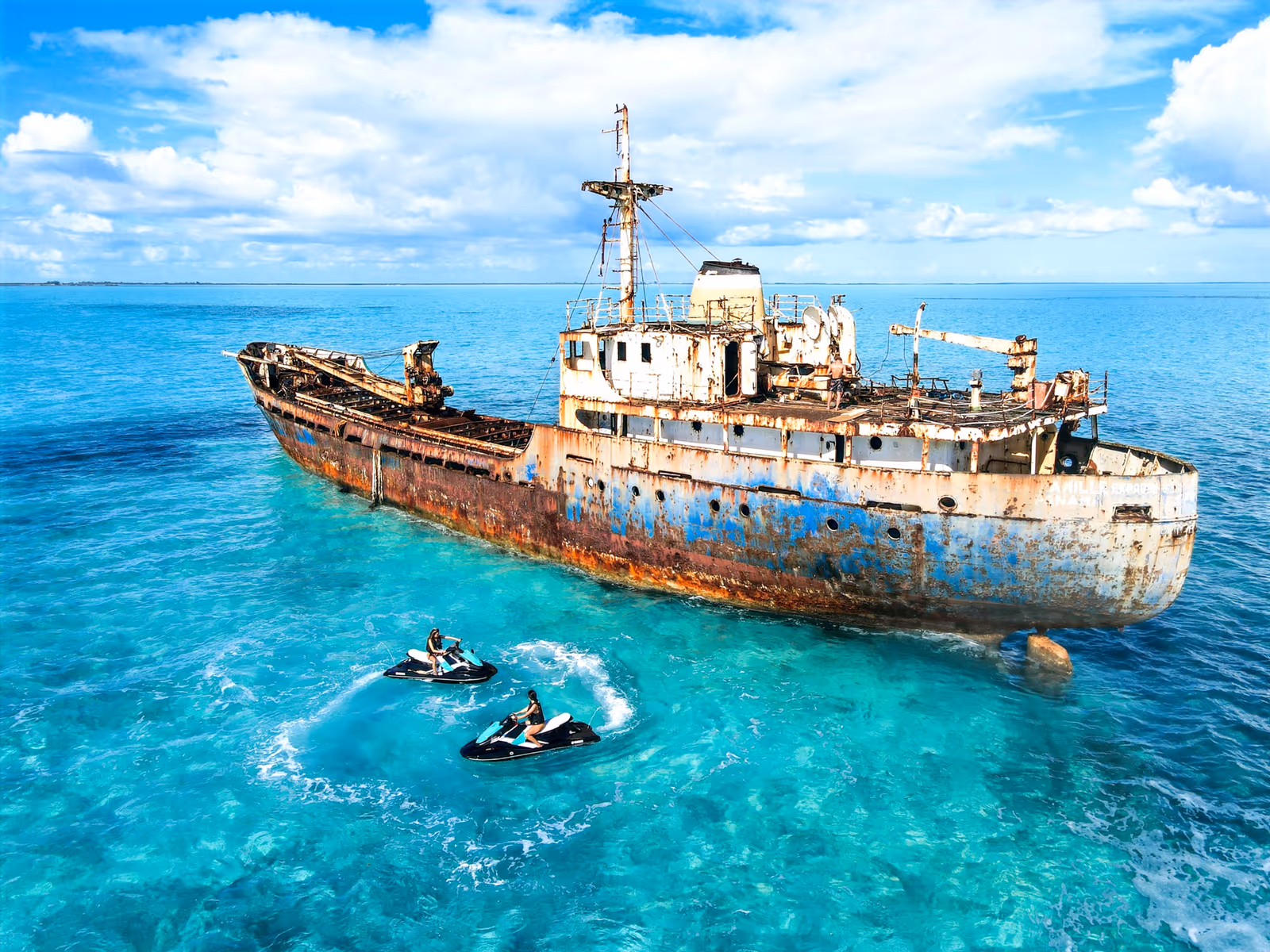 Aerial view of two Jet Skis in clear water beside the rusted hull of a grounded freighter